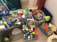 Photo showing multiple plastic bins filled with assorted household and cleaning supply bottles, sponges, cloths, and boxed detergent products including Tide and Pine-Sol.