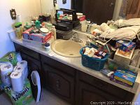Wide view of bathroom counter showing various bottles of lotions, creams, and beauty products, along with packs of paper towels and toilet paper underneath.