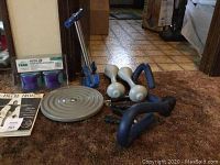 Wide view of exercise equipment on carpet including dumbbells, twist disc, push-up bars, hand exerciser, ankle weights in box, and exercise booklets