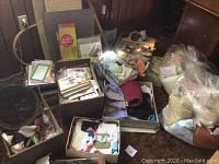 Wide view of boxes containing sewing patterns, fabric pieces, measuring boards, sewing accessories, and craft items in a cluttered setting.