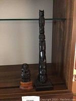 Two Inuit style wood carved sculptures displayed on a wooden shelf under glass, including a tall totem with animal top figure and a smaller seated figure on wooden base.