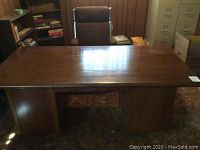 Solid wood desk with polished top surface and visible wood grain, situated in a room with wooden shelves and filing cabinets.