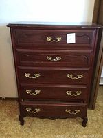 Front view of tall wooden dresser with five drawers, brass handles, and carved decorative details on drawer fronts and legs.