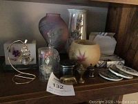 Shelf display showing a variety of small collectibles including a hanging glass angel ornament on a metal stand, decorative glass dome with figurine, ceramic candle holder and vase, small metal containers, and coasters.