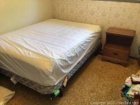 View of double bed with fitted sheet covering mattress, boxspring visible below, and wooden nightstand to side.