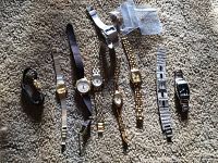 Nine ladies' wristwatches displayed on carpet, showing various bands, face shapes, and colors. Two small clear plastic bags with watch parts appear in view.