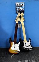 Photo showing two Fender Stratocaster guitar controllers placed standing against a blue wall with PlayStation 3 Rock Band 2 game case above them.