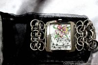 Close-up view of watch face showing square stainless steel bezel and floral orchid design on white dial with chain link bracelet.