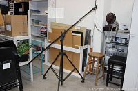 Trimax Light Arm Pro studio boom mounted on a wheeled tripod stand angled upward in a room with shelves and chairs.