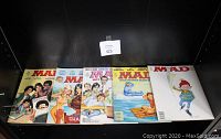 Five MAD magazines displayed side by side showing front covers, showcasing various satirical illustrations and titles from 1977 to 1990.