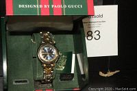 Front view photo showing the Paolo Gucci watch inside its original branded box with green and red stripes. The watch features a deep blue dial and two-tone metal band.