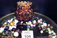 Orange glass vase with attached raised glass marble-like beads surrounded by and filled with an assortment of vintage marbles of various shapes, sizes, and colors on a dark surface with lot number 104 displayed in front.