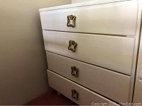 White wooden dresser with four drawers featuring vintage-style brass handles. Shown from left front angle, placed against wall on carpet. Scratches and marks visible on surface.