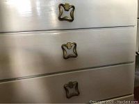 Close-up showing three brass handles on white dresser drawers, detailing hardware shape and finish.