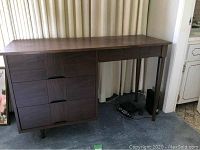Front side view of a wooden desk with three drawers on left side, flat work surface on top, and one leg supporting the right side.