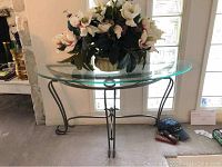 Full view of half moon shaped glass top table with wrought iron base, placed in front of a window, displaying the entire structure and flower arrangement on top.