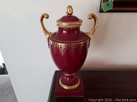 Full visual of red and gold urn with ornate handles and lid finial on wooden surface.