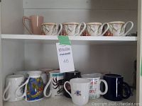 Photo of two shelves showing total 11 ceramic mugs and cups, four white dessert/cappuccino style cups with pink and green leaf and berry design, one tall pink mug, and six assorted mugs with varied designs and colors.