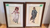 Wide view of the pair of framed needlepoint art pieces: a woman in a white dress and a man in blue clothing.