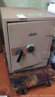 Front view of beige Schwab safe with combination dial and lever, sitting on a worn furniture dolly.
