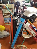 Photo showing two golf bags with clubs on wheeled stands, one red and white, one black, in a cluttered indoor space.