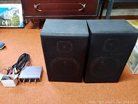 Two black speakers with fabric fronts, showing speaker outlines, and a small metal remote speaker switch with cables beside them on a brown carpeted floor.