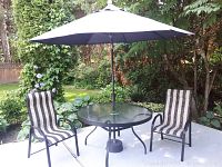 Patio scene showing round glass top table with black metal frame and white umbrella mounted in center, two striped fabric and black metal framed outdoor chairs visible but not included in sale.