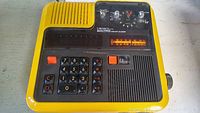 Front and top angled view showing numeric keypad, calculator display, radio tuning scale, speaker, and clock dials on yellow plastic casing.