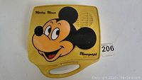 Front view of yellow plastic carrying case for Mickey Mouse phonograph with a large colorful graphic of Mickey Mouse's face, built-in speaker holes on the right side, and 'Mickey Mouse Phonograph' text.