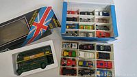 Top view of 24 Hot Wheels cars arranged in blue plastic storage case with compartments beside original packaging for larger green London double-decker bus.