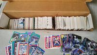 Open cardboard box filled with sports trading cards standing vertically packed tightly inside. Baseball and hockey cards laid out in front of box showing various designs and colors.