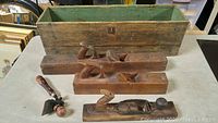 Photo of lot showing two wooden hand planes, a metal hand plane, a paintbrush with wooden handle, and a large wooden box with green interior.