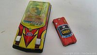 Two vintage tin toy cars on a white surface; one larger colorful car with transparent roof, one smaller red fire department car labeled 'CHIEF'.