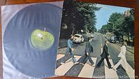 Vinyl record partially in inner plastic sleeve with visible yellow-green Apple label, next to album cover showing Beatles crossing Abbey Road.