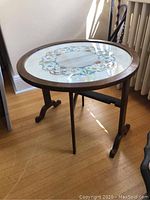 Full view of round wooden needlepoint side table with glass top, tripod legs, and fold-down feature.