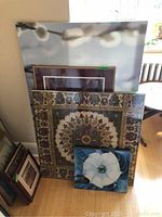 Three framed prints stacked against a wall showing different styles: floral, mandala, and blue flower. Various frame sizes visible.