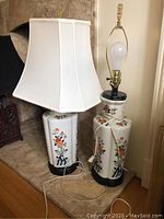 Two porcelain table lamps placed on a mantel. One lamp has a white fabric shade; the other does not have a shade. Both lamps have floral painted decoration and black bases.