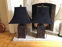 Pair of two table lamps with carved dark wood bases and black square lampshades placed on wooden surface in front of fireplace.