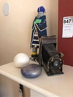 Wide photo showing the vintage folding camera, two stone paperweights and beaded statue on a white shelf against a red wall.