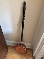 Full view of the copper food warmer with long wooden handle leaning against a wall on a hardwood floor.