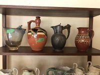 Four decorative pitchers on a shelf: glass, ceramic with multicolor glaze, metal with lid, and ceramic with raised geometric pattern