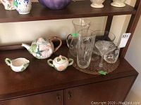 Photo of shelf containing ceramic tea pot with floral and bird design, matching sugar and creamer bowls, and various clear glass bar ware including pitchers and glasses