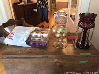Overview of the lot showing clear antique glass candy container filled with gold Christmas balls, cranberry glass lamp with hanging prisms, and packaged purple and gold Christmas balls.