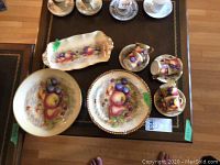 Overview of all fruit motif china pieces arranged on table showing cups, saucers, plates, platter and bowl