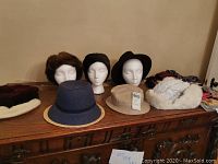 Photo showing six hats placed on and beside mannequin heads on a wooden surface: brown fur hat, black textured hat, black felt Tiley Fedora, blue woven straw hat with tan rim, tan bucket-style hat, white fur-trimmed winter hat. Scarves seen folded at side.