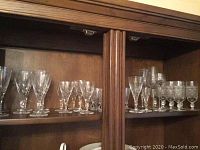 Wide view of wooden cabinet shelf displaying crystal and glass wine glasses with varied stem decorations including leaf and twisted designs