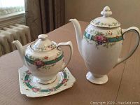 Two bone china Royal Doulton tea and coffee pots with floral turquoise pattern and gold trim on a wooden table by a window.