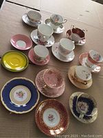 Photo displaying the assorted tea cups and saucers arranged on cardboard surface, showing various colors, floral designs, and gold trim details.