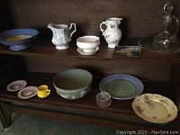 Shelved collection showing various pitchers, bowls, plates, and a glass decanter with stopper.