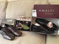 Two pairs of shoes side by side with three clutch purses behind them on a sofa, including Amalfi black ladies shoes in box.